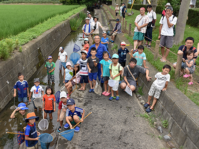 写真：用水路で天野　武雄さんを囲むたくさんの子供たち